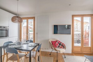 Une cuisine moderne équipée d’un four et d’un micro-ondes encastrés, un coin repas avec une table pour quatre personnes, et un espace salon confortable avec un fauteuil et un téléviseur fixé au mur.