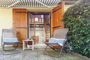 Un espace extérieur abrité, avec deux chaises et une petite table, une porte en bois et une haie.