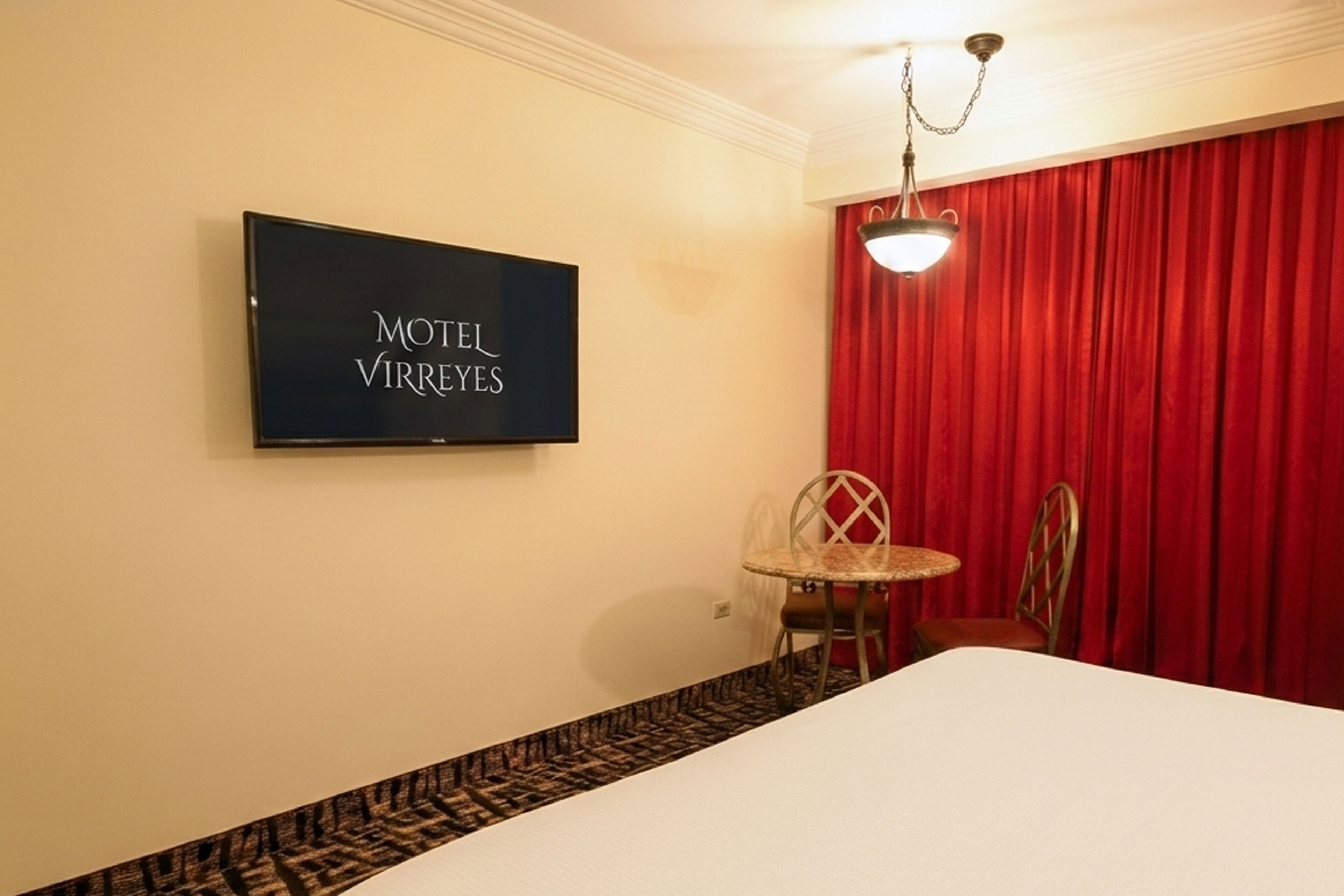 Een hotelkamer met een bed, een televisie waarop 'MOTEL VIRREYES' staat, een rood gordijn en een kleine tafel met stoelen.
