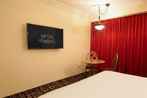 Habitación de hotel con una cama, un televisor mostrando “MOTEL VIRREYES”, una cortina roja y una pequeña mesa con sillas.