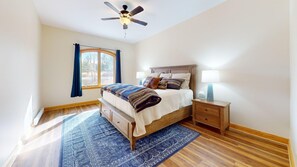 Une chambre à coucher comprenant un lit, des tables de chevet, un ventilateur de plafond, une fenêtre avec des rideaux et un tapis à motifs.