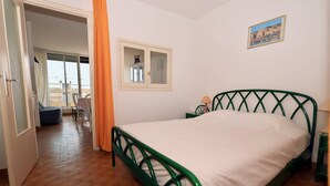 Ein Schlafzimmer mit einem grünem Metallbettgestell, einer weißen Matratze, einem kleinen Nachttisch mit Lampe und einem Fenster mit Bilderrahmen.