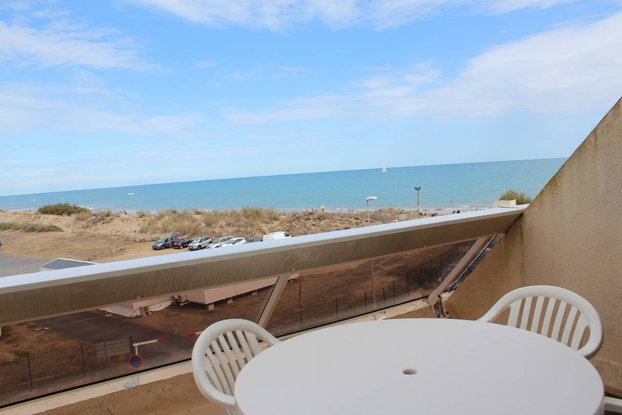 A balcony with a table and chairs overlooking a beach with parked cars and a sailboat in the distance.