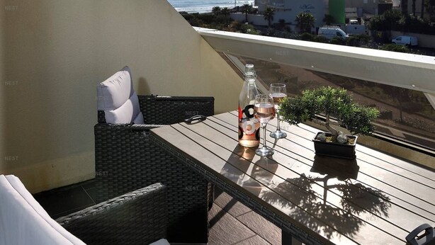 Ein Balkon mit einem Korbstuhl, einem kleinen Tisch, einer Flasche und zwei Gläsern. Im Hintergrund ist ein Gewässer und Gebäude zu sehen.