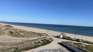 Une vue sur la plage, avec un ciel dégagé, un sentier sablonneux menant à la mer, et quelques personnes au loin.