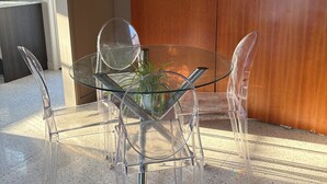 Een tafel met glazen blad, vier transparante stoelen, een potplant en een houten garderobe in een moderne kamer met een betegelde vloer.