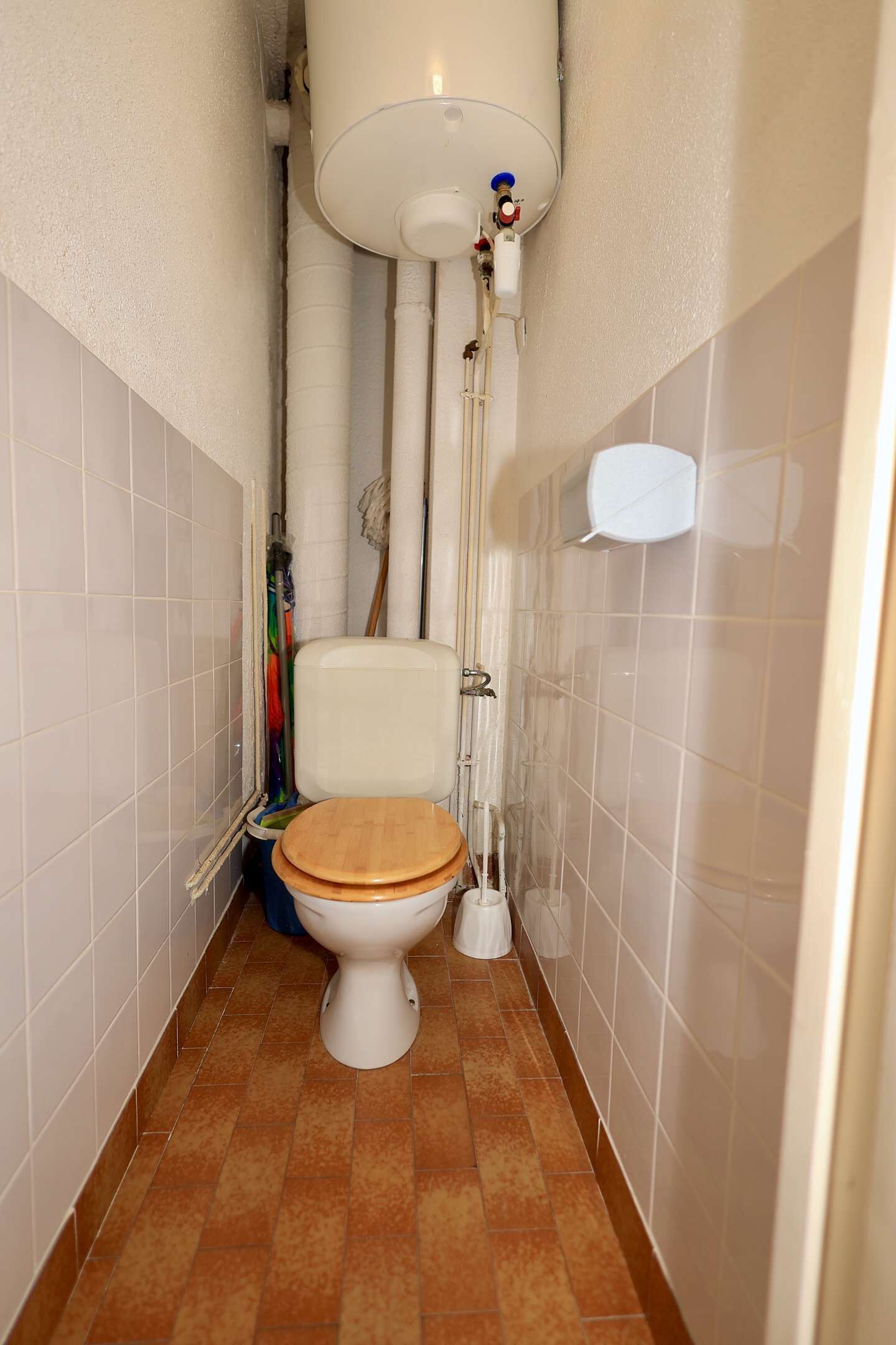 A small bathroom with a toilet, white tiles, and a brown floor.