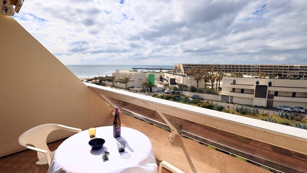 Un balcón con una mesa, sillas y una botella de vino. Se ve una playa, palmeras y un edificio de varios pisos.