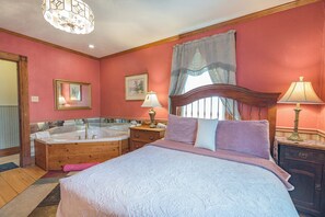 A bedroom with a large bed, a wooden side table, a nightstand, a mirror, a window with curtains, and a bathtub.