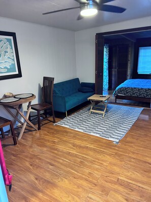 Una habitación con un sofá azul, una pequeña mesa de madera, una silla, una cama con cubrecama azul y un ventilador de techo.