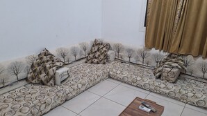 Ein Wohnzimmer mit einer Eckcouch, einem kleinen Tisch mit Fernbedienungen und einem Fenster mit Vorhängen.