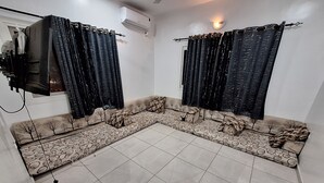 Ein Wohnzimmer mit Ecksofa, zwei Fenstern mit Vorhängen, einem flachbildschirmfernseher an der Wand und einem gefliesten Boden.