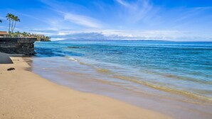 Una playa con agua azul cristalina, una orilla de arena y un edificio con palmeras a la izquierda.