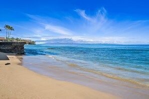A beach with clear blue water, a sandy shore, and a building with palm trees on the left. - SUMMER SALE! Direct Oceanfront 2BR with Superb Views  - Nohonani 401 (Lahaina)