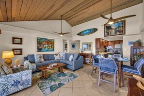 A spacious living room with a blue sofa, a dining area with wicker chairs, and a kitchen in the background. - SUMMER SALE! Direct Oceanfront 2BR with Superb Views  - Nohonani 401 (Lahaina)