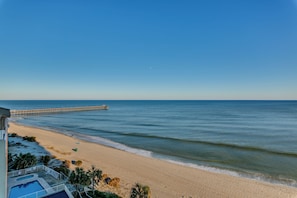 Blick auf einen Strand mit blauem Himmel, sandigem Ufer und einem Steg, der sich ins Wasser erstreckt.