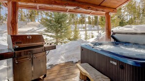 Un terrazzo in legno con una vasca idromassaggio e un barbecue, circondato da neve e alberi.