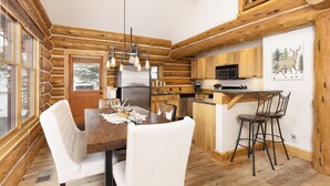 Interno di una baita in legno con un tavolo da pranzo in legno, sedie bianche, una cucina con armadietti in legno, un forno a microonde e un’area bar con sgabelli.