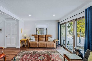 Ein gemütliches Wohnzimmer mit einem braunen Ledersofa, einem gemusterten Teppich und einem großen Fenster mit Blick ins Freie.