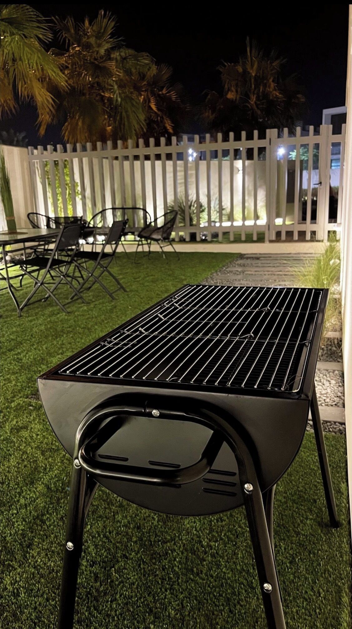 Ein schwarzer Grill mit einem Metallrost, auf einer Rasenfläche mit weißem Zaun und Palmen im Hintergrund.