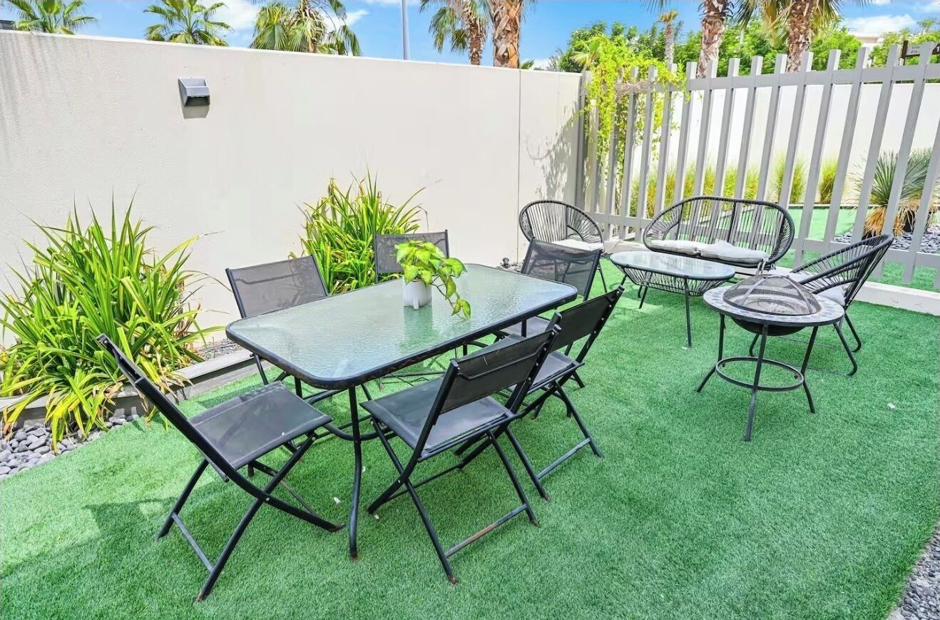 Um pátio externo com mesa de vidro e cadeiras de metal preto, rodeado por grama sintética verde e plantas.