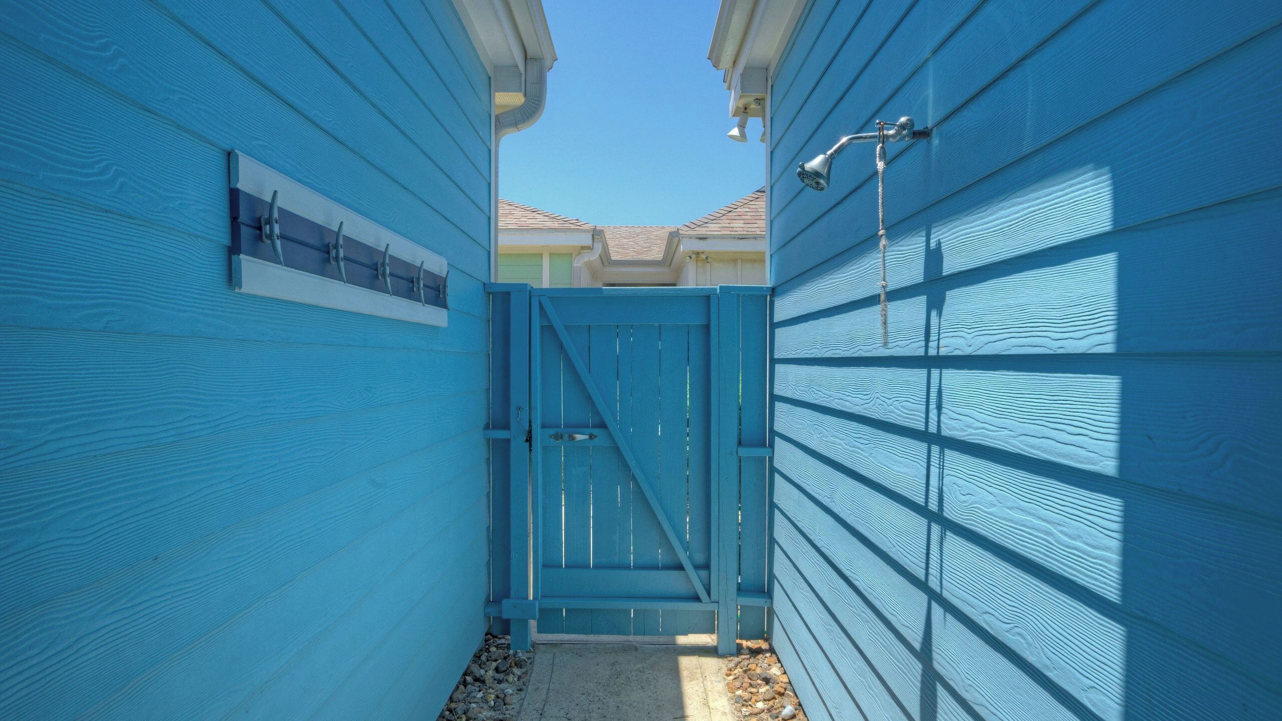 A narrow pathway between two blue-sided buildings with a metal gate and a water spout on the right.