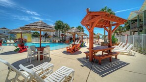 A pool area with lounge chairs, a wooden pergola, and thatched umbrellas. - The Blue Pelican by Avantstay Pirates Bay Retreat + 2 Shared Lagoon Pools 3 Blocks to Beach (Port Aransas)