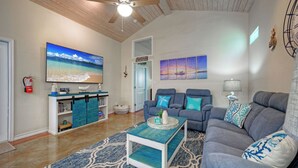 A living room with a large TV, two sofas, a coffee table, and a bookshelf. - The Blue Pelican by Avantstay Pirates Bay Retreat + 2 Shared Lagoon Pools 3 Blocks to Beach (Port Aransas)