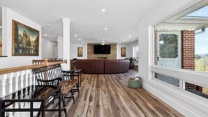Un salón espacioso con piso de madera, una mesa de comedor con sillas, un televisor grande montado en una pared de piedra y vistas a través de una ventana.