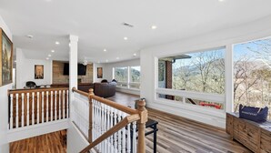 Un amplio salón con terraza de madera, ventanas grandes que ofrecen vista a árboles, y una decoración interior moderna.