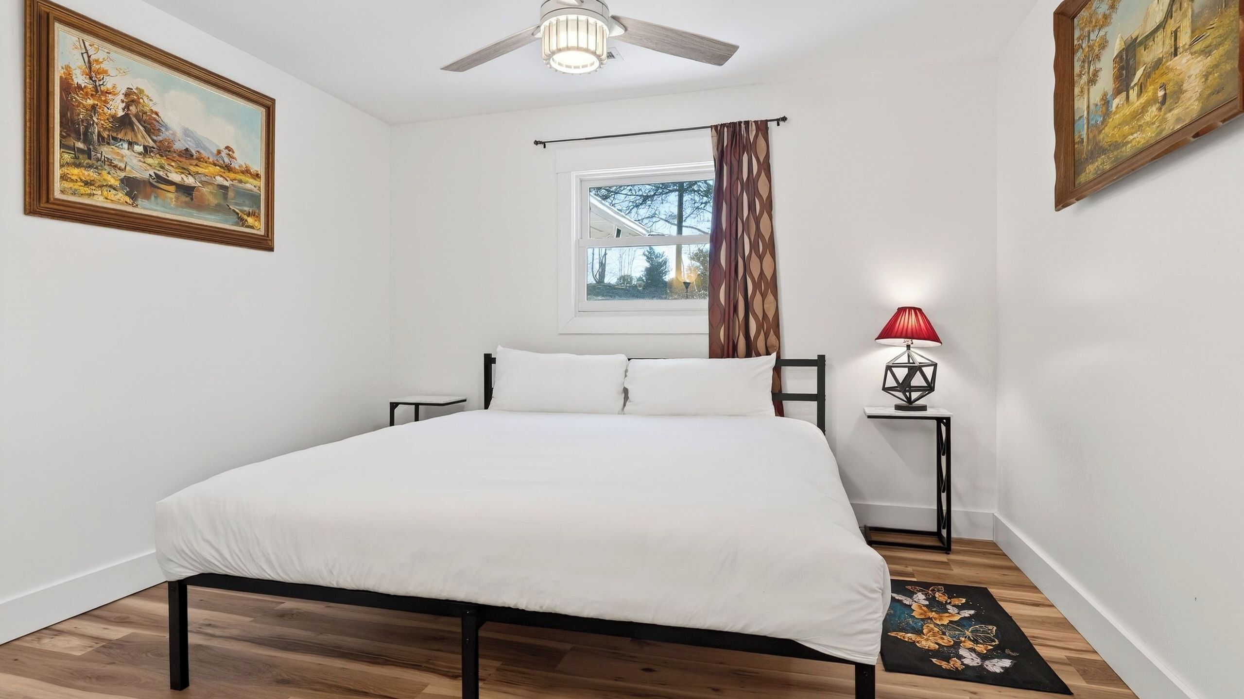 A bedroom with a bed, a ceiling fan, a window with curtains, a nightstand with a lamp, and two paintings on the walls.
