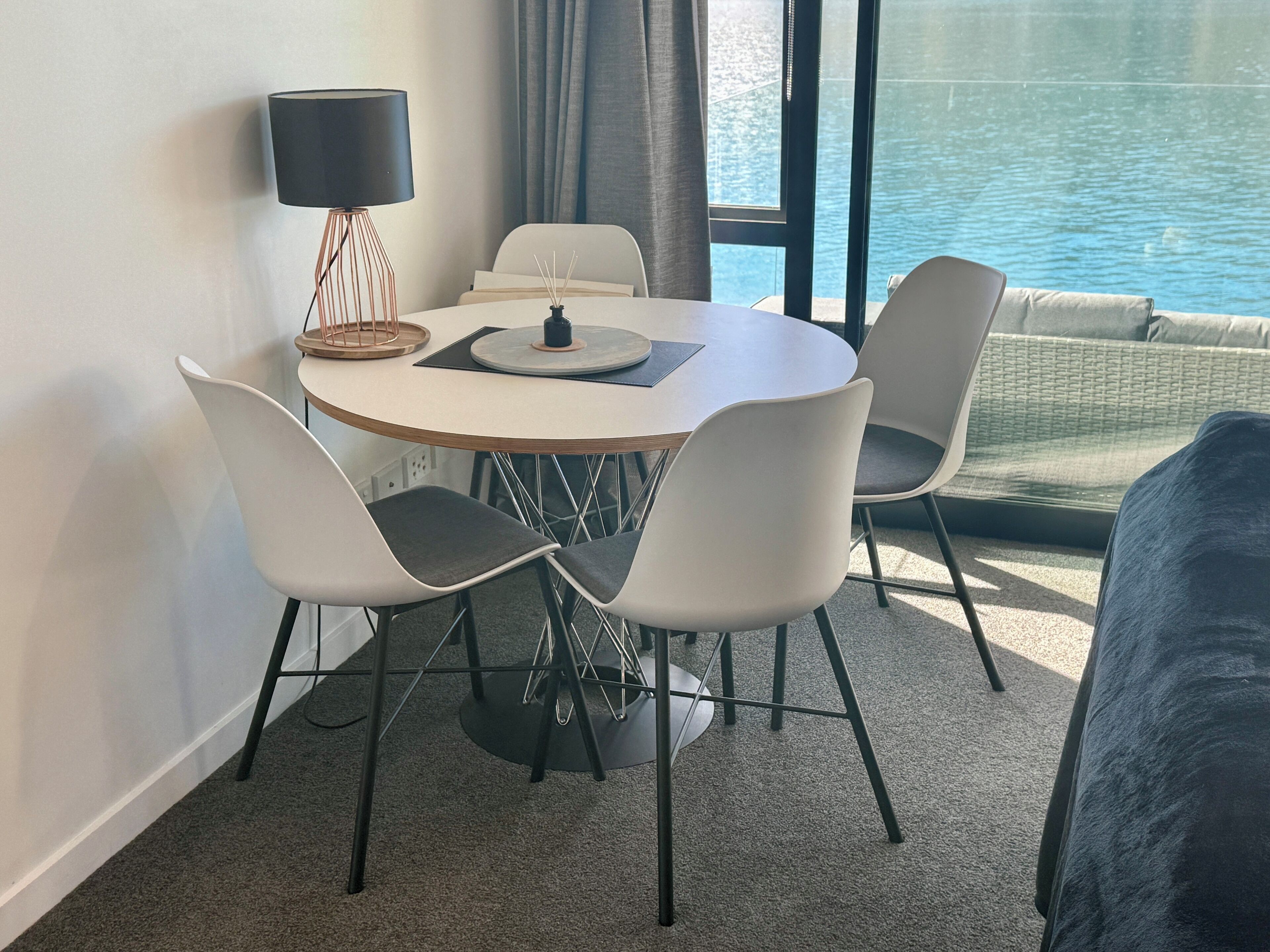 A modern dining area with a round table, four chairs, a lamp, and a view of the water.