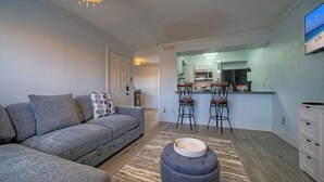 A living room with a grey sofa, a patterned throw pillow, a round ottoman, and a kitchen area with a bar stool.