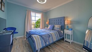 A bedroom with a bed, a desk, a chair, a window with curtains, and a nightstand with a lamp.