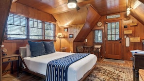 A cozy wooden cabin bedroom with a bed, a desk, and a chair. There are windows with blinds, a clock, and a sign labeled 'antiques'.