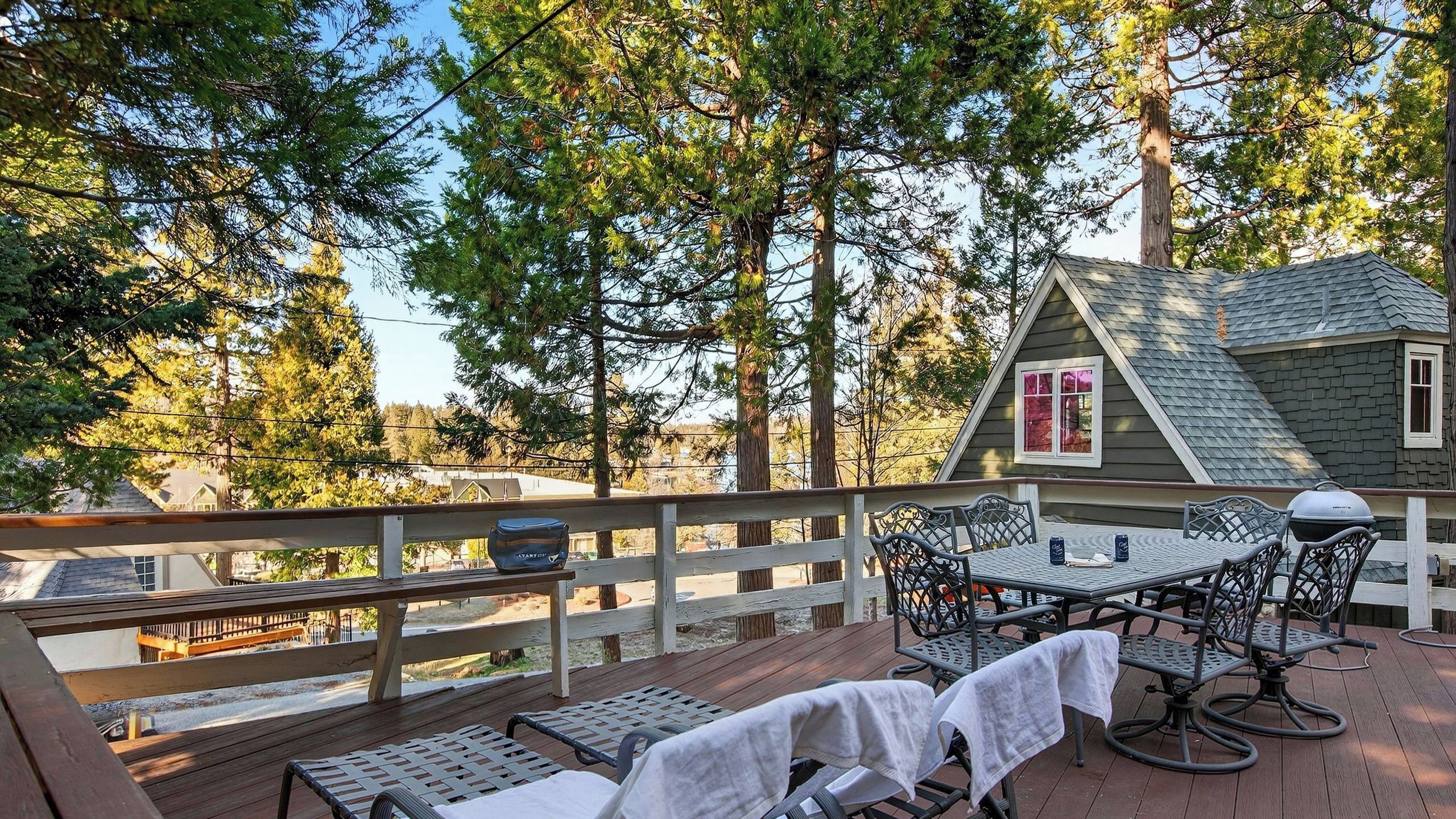 A wooden deck with outdoor furniture, a small house with a red window, and tall trees in the background.
