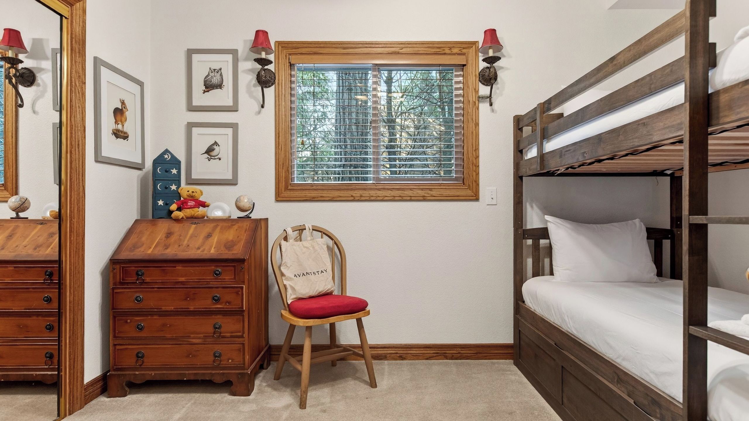 Une chambre avec un lit superposé, une commode, une chaise avec un coussin rouge et une fenêtre avec des stores.