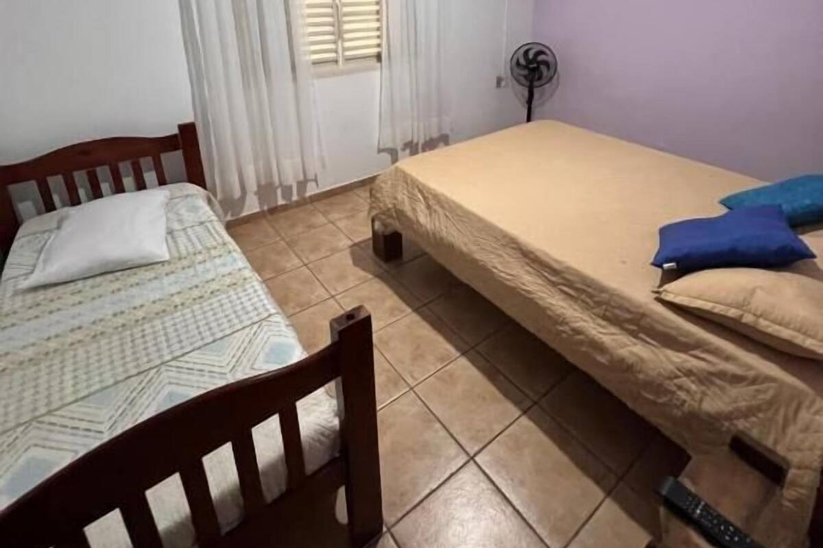 Une petite chambre bien éclairée, avec un lit simple, une chaise en bois, un ventilateur de plafond et une fenêtre avec des rideaux.