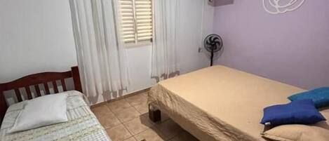 A small, well-lit bedroom with a single bed, a wooden chair, a ceiling fan, and a window with curtains.