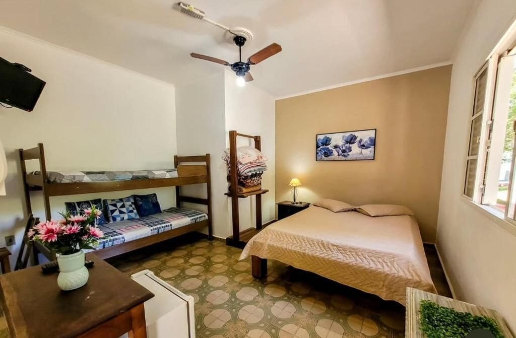 A compact room with a bunk bed, a single bed, a ceiling fan, a TV mounted on the wall, and a window with a view.