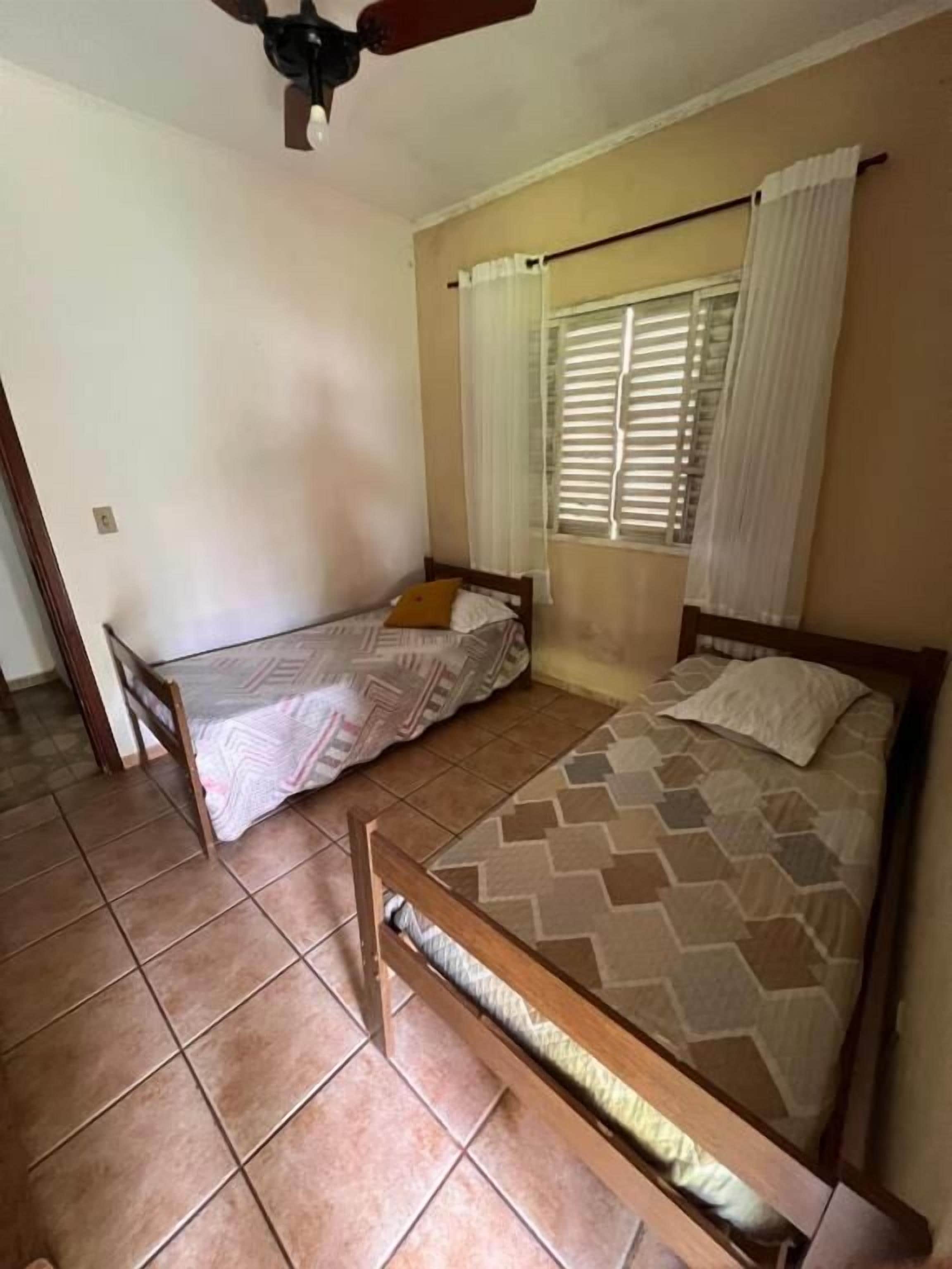 Une chambre avec deux lits simples, un ventilateur de plafond, une fenêtre avec des stores et un sol carrelé.