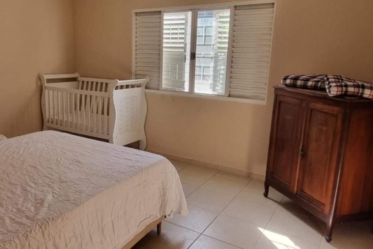 Une chambre à coucher avec un lit, un berceau, une armoire en bois et une fenêtre avec des stores.