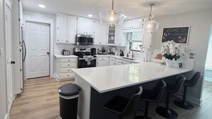 A modern kitchen with white cabinets, a large island with bar stools, a black trash bin, and a chalkboard sign. - CASABELLA,4bed/2bath, 8 min from beach/ w/ POOL (Fort Walton Beach)