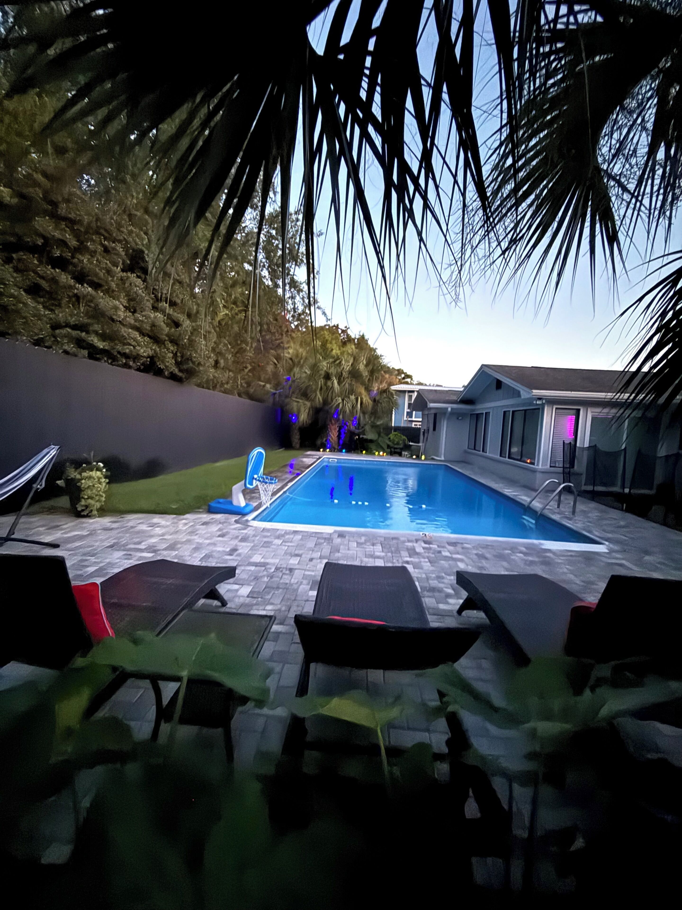 Una piscina con sillas de descanso, un aro de baloncesto y una casa al fondo.