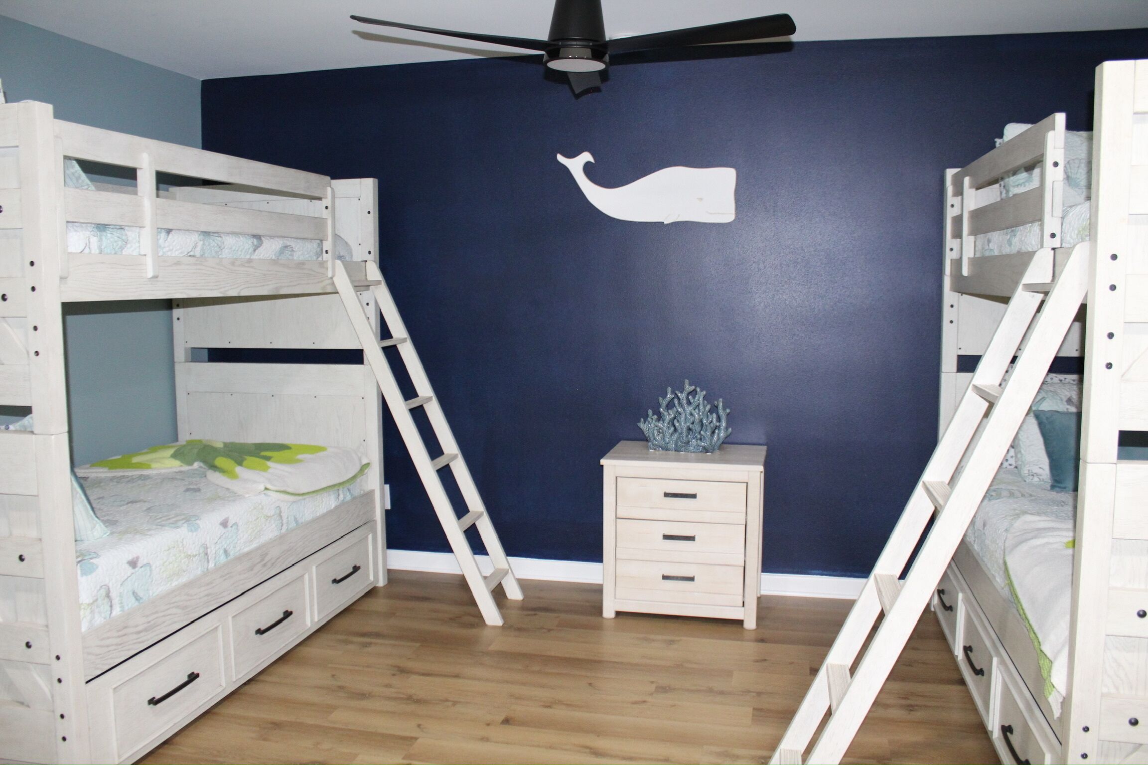 A room with bunk beds, a ceiling fan, a small bedside table, and a wall decoration.