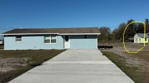 A single-story house with a light blue exterior and a gray roof. There is a concrete driveway leading to the front door, which has a small porch area. The house is surrounded by a grassy yard with a dirt path to the right. - Modern Farmhouse Quiet & Spacious located in Safe neighborhood-RV parking space (Riverview)