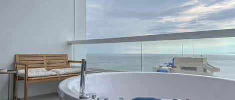A balcony with a wooden chair, a white bathtub, and a view of the sea.
