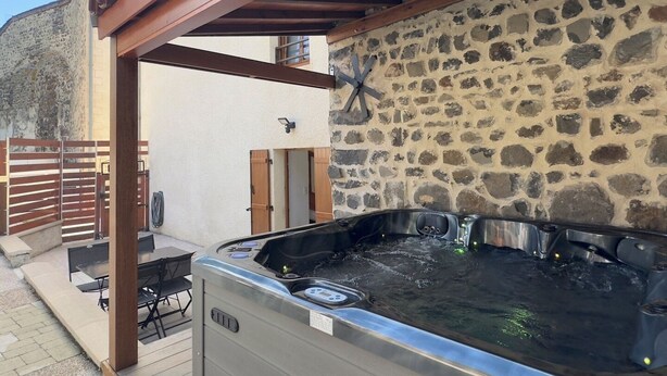 Un bain à remous sous une pergola en bois avec des murs en pierre, une table et des chaises à l’extérieur, et un portail.