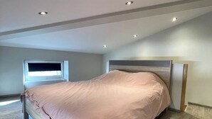Een slaapkamer met een bed, houten vloeren, een raam en inbouwspots.