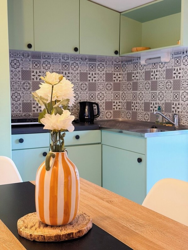 Une cuisine avec des meubles de couleur vert clair, un pare-feu décoré de motifs et une table en bois sur laquelle est posé un vase rayé rempli de fleurs blanches.